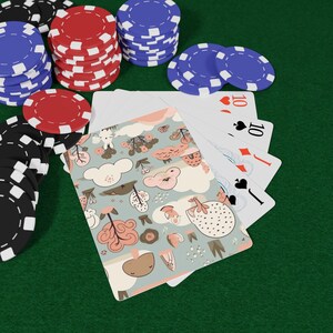 Può includere: Un mazzo di carte da gioco con il dorso azzurro decorato da un motivo di alberi rosa, nuvole bianche e piccoli animali, disposto a ventaglio per mostrare dieci di cuori, dieci di picche, fante di quadri e fante di picche, tra pile di fiches.