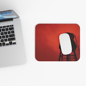 May include: A rectangular mouse pad featuring a silhouette of a water tower against a vibrant red sky. A white computer mouse rests on the surface of the mouse pad, which is positioned next to a silver laptop with a black keyboard and screen.