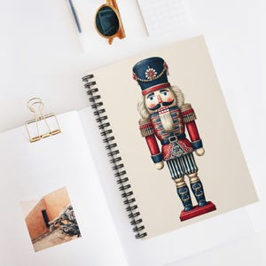 Puede incluir: Cuaderno de espiral con una ilustración colorida de un soldado cascanueces tradicional. El cascanueces está representado con un uniforme rojo y azul con detalles dorados. El cuaderno está abierto, con un clip dorado y gafas de sol cerca.