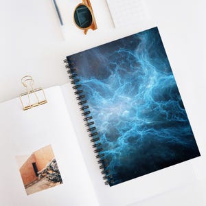 Op de afbeelding: Spiraalgebonden notitieboek met een levendig blauw en wit nevelontwerp op de omslag. Het notitieboek is open en toont blanco witte pagina's. Een goudkleurige paperclip en zonnebril zijn ook zichtbaar.