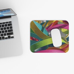 Peut inclure: Tapis de souris rectangulaire avec un motif abstrait coloré de rubans superposés dans des tons or, turquoise, vert, rose et jaune. Une souris d'ordinateur blanche repose sur le tapis. Un ordinateur portable est partiellement visible en haut à gauche.