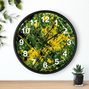 May include: Round wall clock with a black frame. The clock face features a vibrant abstract design with yellow, green, black, and white paint splatters. White numerals mark the hours, and black hands indicate the time.