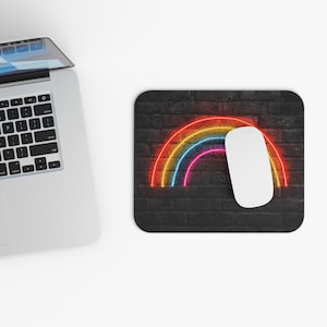 May include: A black rectangular mousepad with a neon rainbow design in red, yellow, blue, and pink. A white computer mouse rests on the mousepad. The background has a brick texture.