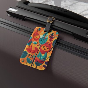 May include: A rectangular luggage tag featuring a vibrant pattern of stylized leaves and faces in shades of orange, red, teal, and yellow. The tag is attached to a dark brown suitcase handle by a black strap with a gold-toned metal buckle.