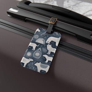 May include: A rectangular luggage tag featuring a dark blue background with a repeating pattern of white polar bears, stylized igloos, and geometric shapes. The tag is attached to a dark brown suitcase handle with a black strap and a gold buckle.