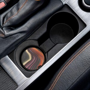 May include: A car cup holder with a colorful abstract design coaster. The coaster features a swirl of orange, pink, and white colors. The cup holder is black and sits in a car's console, surrounded by black leather and silver trim.