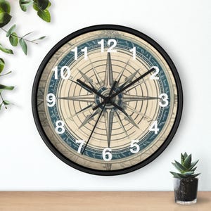Op de afbeelding: Een ronde wandklok met een zwarte rand en een beige wijzerplaat met een vintage kompasroosontwerp. De klok heeft dikke witte cijfers van 1 tot 12 en dunne zwarte wijzers tegen een achtergrond met concentrische blauwe cirkels en lijnen.