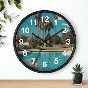 May include: A round black wall clock featuring a scenic image of a swimming pool with palm trees under a blue sky. The clock has white numerals and black hands, with a small succulent plant in a pot on the right.