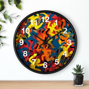 May include: A round wall clock with a black frame. The clock face features a colorful abstract design with yellow, blue, orange, and red figures. White numerals mark the hours. The clock is on a wooden surface.