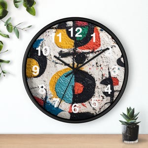 May include: A round wall clock with a black frame. The clock face features an abstract design with white, yellow, blue, red, and black shapes. White numbers mark the hours. The clock hands are black.