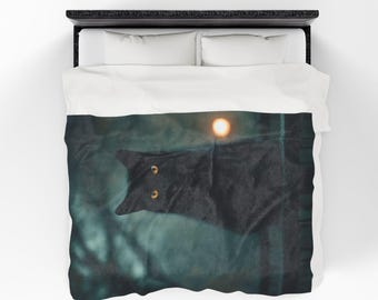May include: A black cat with yellow eyes is featured on a duvet cover. The cat is positioned in the center of the image, with a blurred background of green and a bright, round light source. The bedding is white and the bed frame is black.