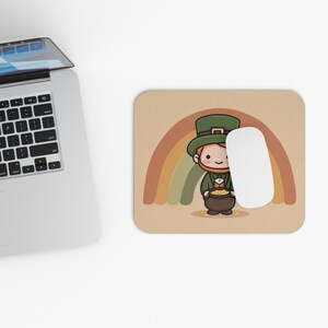 May include: A beige mouse mat with a cartoon leprechaun holding a pot of gold, with a rainbow above. The leprechaun wears a green hat and coat. A white computer mouse rests on the mat, next to a laptop.