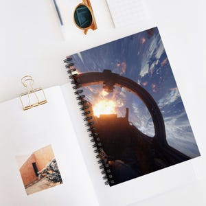 May include: Spiral-bound notebook featuring a dramatic image of a cockpit view with a fiery explosion against a backdrop of Earth and space. The notebook is open, revealing a photograph and a gold paper clip.