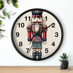 Könnte beinhalten: Eine runde Wanduhr mit schwarzem Rahmen. Das Zifferblatt zeigt eine detaillierte Illustration eines Nussknacker-Soldaten in Rot, Blau und Gold. Die Zeiger sind schwarz, und die Zahlen sind in einer klassischen Schriftart.