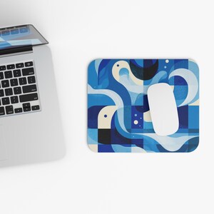 May include: A rectangular mousepad with a blue abstract design featuring white and black shapes. A white wireless mouse rests on the pad, next to a laptop. The mousepad has a geometric pattern with a modern aesthetic.