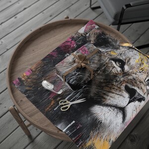 May include: A wrapped gift and wrapping paper featuring a lion's face with a brown bow. The wrapping paper has a black background with pink, yellow, and white accents. Scissors and tape are also on the wooden table.