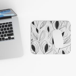 May include: A white mousepad with a black and white floral design and a white computer mouse. The floral pattern features stylized leaves and seed pods. A silver laptop is partially visible in the upper left corner.