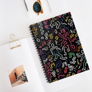 Puede incluir: Un cuaderno negro con espiral con un diseño vibrante de figuras danzantes y notas musicales en blanco, rosa, amarillo, verde y azul. El cuaderno está abierto sobre una superficie blanca, con un clip dorado y gafas de sol visibles.
