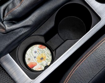 Könnte beinhalten: Ein Getränkeuntersetzer für den Auto-Getränkehalter mit einem Blumendesign mit gelben, orangefarbenen und weißen Blumen. Der Untersetzer ist rund und passt in den Getränkehalter eines Autos. Der Innenraum des Autos ist schwarz und silberfarben.