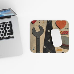 Könnte beinhalten: Rechteckiges Mauspad mit beigem Hintergrund mit einem grauen Schraubenschlüssel, einer schwarzen Krawatte mit weißen Punkten und einem orangefarbenen Herz. Eine weiße Computermaus liegt auf dem Pad, in der Nähe eines Laptops.