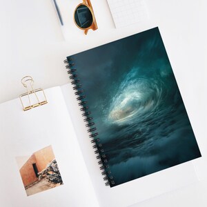 Puede incluir: Cuaderno de espiral con un llamativo diseño de ola oceánica en tonos turquesa y blanco. El cuaderno está abierto, mostrando una página en blanco y un clip dorado. También se ven gafas de sol y un bolígrafo.