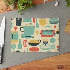 May include: A rectangular glass cutting board featuring a cream-colored background with a repeating pattern of kitchen appliances and utensils in teal, yellow, red, and dark blue, including a stand mixer, toaster, spatula, and various bowls.