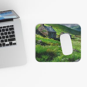 May include: Mouse mat with a watercolour painting of a stone cottage in a green, grassy landscape with rolling hills. A white computer mouse sits on the mat near a laptop.