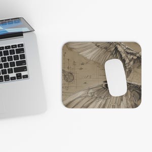 May include: A computer mouse and mousepad featuring a sepia-toned illustration of a bird's wings and architectural blueprints. The mousepad has a white oval mouse on top. A laptop is in the upper left corner.