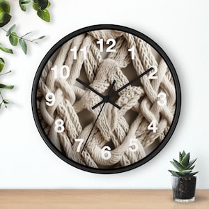 May include: A round wall clock with a black frame and a face featuring a beige macrame cord pattern. The clock has white Arabic numerals from 1 to 12 and three black hands indicating the time. The design displays a textured, knotted rope aesthetic.