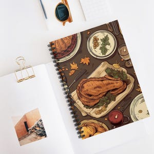May include: Spiral-bound notebook with a Thanksgiving dinner illustration. The cover shows a roasted turkey, side dishes, and autumn leaves on a brown background. The notebook is open on a white surface.