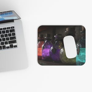 May include: A computer mouse rests on a mousepad featuring a row of colorful potion bottles. The bottles are filled with orange, purple, yellow, and teal liquids, each with a cork stopper and a chain. The background is dark.