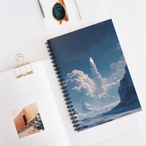 May include: Spiral-bound notebook featuring a vibrant image of a rocket launching into a cloudy, blue sky. The notebook's cover showcases a detailed illustration of a rocket blasting off, with white clouds and a snowy landscape below. The notebook is open, revealing blank pages.