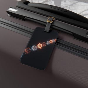 May include: A rectangular black luggage tag attached to a dark brown suitcase handle with a black strap and a gold-toned buckle. The tag features a diagonal graphic design of glowing, fiery circular orbs in shades of orange, red, and white.