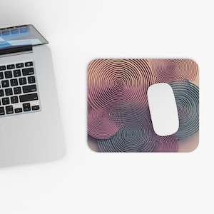 May include: A rectangular mouse pad featuring a pattern of overlapping concentric circles in shades of pink, purple, and blue. A white computer mouse rests on the surface of the pad, which is positioned next to a silver laptop with a black keyboard.
