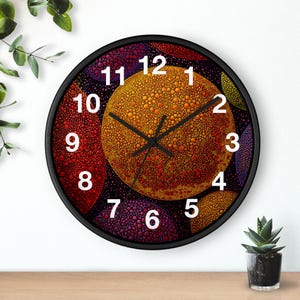 May include: A round wall clock with a black frame. The clock face features a colorful abstract design with orange, red, and purple circular patterns. White numbers mark the hours, and black hands indicate the time.