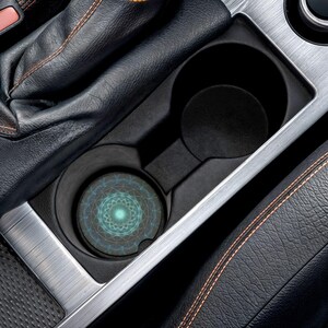 May include: A car cup holder with a decorative coaster featuring a teal and blue geometric design. The coaster is placed in the front cup holder of a vehicle, surrounded by black leather and silver trim.