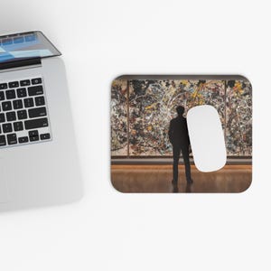 May include: A rectangular mousepad featuring a person silhouetted in front of a large abstract painting. A white computer mouse is positioned to the right of the painting. The painting has a colorful splatter design.