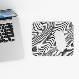 May include: A rectangular mousepad with a white oval cutout for the mouse. The mousepad features a black and white wavy pattern. A white computer mouse is in the cutout. A laptop is in the background.