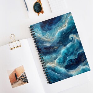 May include: Spiral-bound notebook featuring a vibrant abstract design of swirling blue and teal colors with gold flecks. The notebook is open on a white surface, with a gold paper clip and sunglasses nearby. The cover design resembles a celestial or oceanic theme.