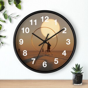 May include: Round wall clock with a black frame. The clock face features a surreal landscape with figures, a large sun, and a golden ring. White numerals mark the hours, and black hands indicate the time.