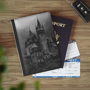 May include: A black and white passport holder featuring a gothic castle image, next to a navy blue passport, two boarding passes, and a car key. The passport holder is made of leather-like material, and the boarding passes have blue and white details.