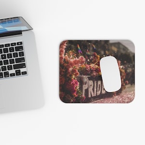 Può includere: Un tappetino per mouse rettangolare con un mouse bianco sopra. Il tappetino presenta una composizione floreale con la parola "PRIDE" a lettere grandi. Una bandiera arcobaleno è visibile sullo sfondo.