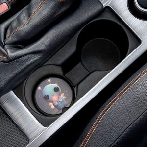 May include: A car coaster featuring a cartoon alien with large black eyes, holding a flower. The coaster is in a car's cup holder, surrounded by black leather and silver trim. The alien is light blue, with pink and yellow accents.