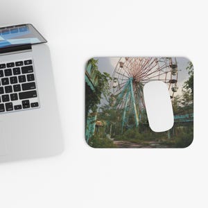 May include: Mousepad featuring a vintage image of a decaying Ferris wheel and overgrown vegetation. The image has a faded, distressed look with muted colors. A white computer mouse rests on the mousepad, next to a laptop.
