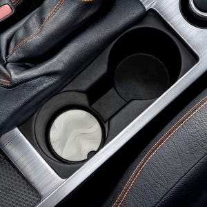 Könnte beinhalten: Ein Getränkehalter im Auto mit einem silbernen Untersetzer mit Wellenmuster. Der Getränkehalter ist schwarz und hat eine Zierleiste aus gebürstetem Metall. Der Innenraum ist aus schwarzem Leder mit orangefarbenen Nähten.
