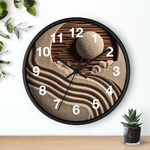 May include: A round wall clock with a black frame and a face featuring a textured sand garden design. The dial displays white Arabic numerals from 1 to 12 and black hour, minute, and second hands. The background shows a raised sphere and raked sand patterns.