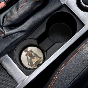 Puede incluir: Un posavasos redondo para coche con una ilustración steampunk de una figura femenina con un vestido. El posavasos está colocado en el portavasos de un coche, rodeado de cuero negro y molduras plateadas.