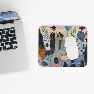 Op de afbeelding: Een rechthoekige muismat met een kleurrijke illustratie van een afstudeerfeest. De scène bevat mensen, een taart, ballonnen en een tafelsetting. Een witte computermuis ligt op de mat, naast een laptop.