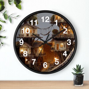 Puede incluir: Un reloj de pared redondo con marco negro que presenta una imagen impresa de varias casas en los árboles iluminadas conectadas por puentes de cuerda. La esfera tiene números blancos grandes del 1 al 12 y manecillas negras.