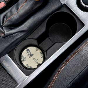 May include: A round car coaster with a science theme, featuring illustrations of a microscope, beakers, and atomic symbols on a grid background. The coaster is placed in a car's cup holder, surrounded by black leather and silver trim.